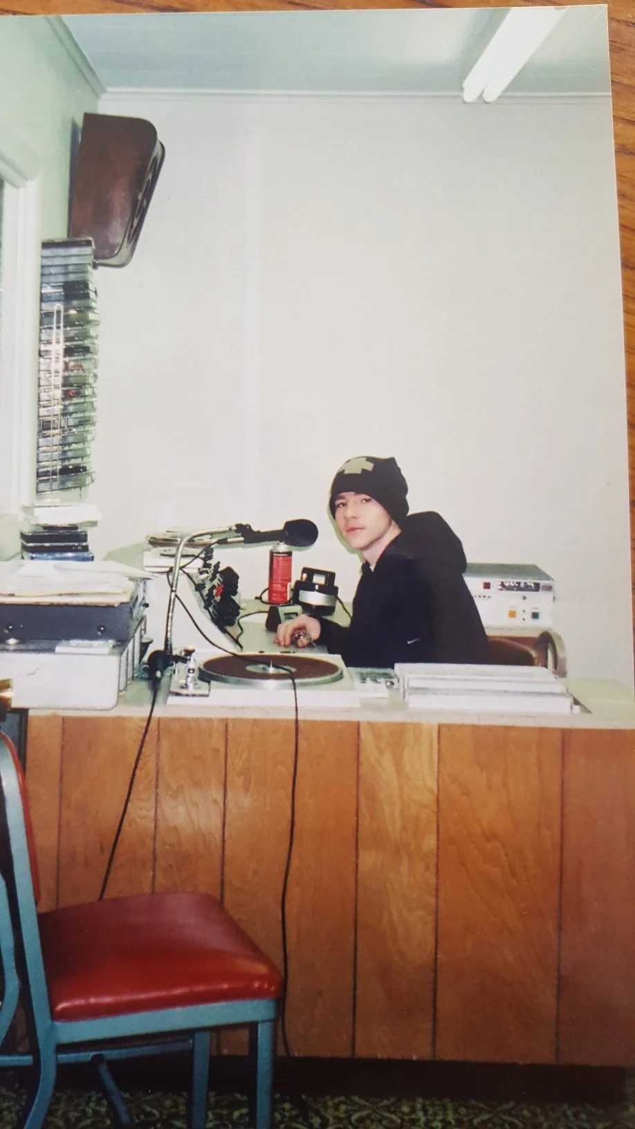 Young man sitting at a radio station desk with a turntable, microphone, and radio equipment. He is wearing a black hoodie and a camouflage beanie, looking at the camera.