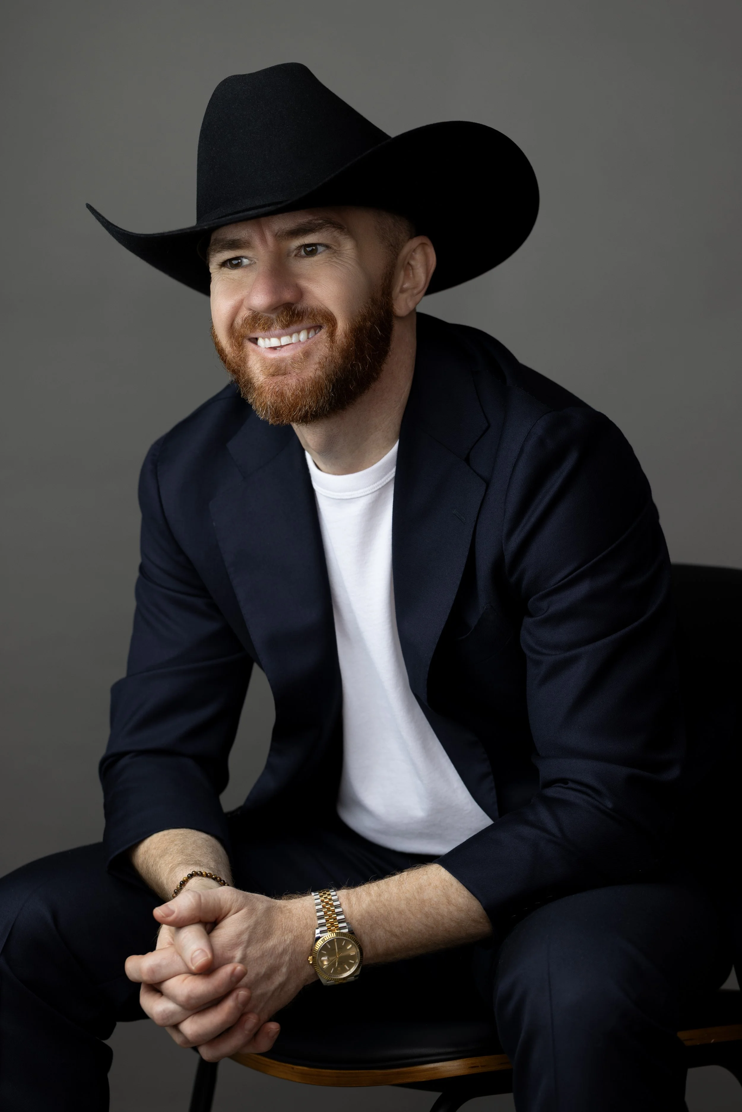 A man with a red beard wearing a black cowboy hat, white T-shirt, and navy blazer seated on a chair against a gray background, smiling with hands clasped.