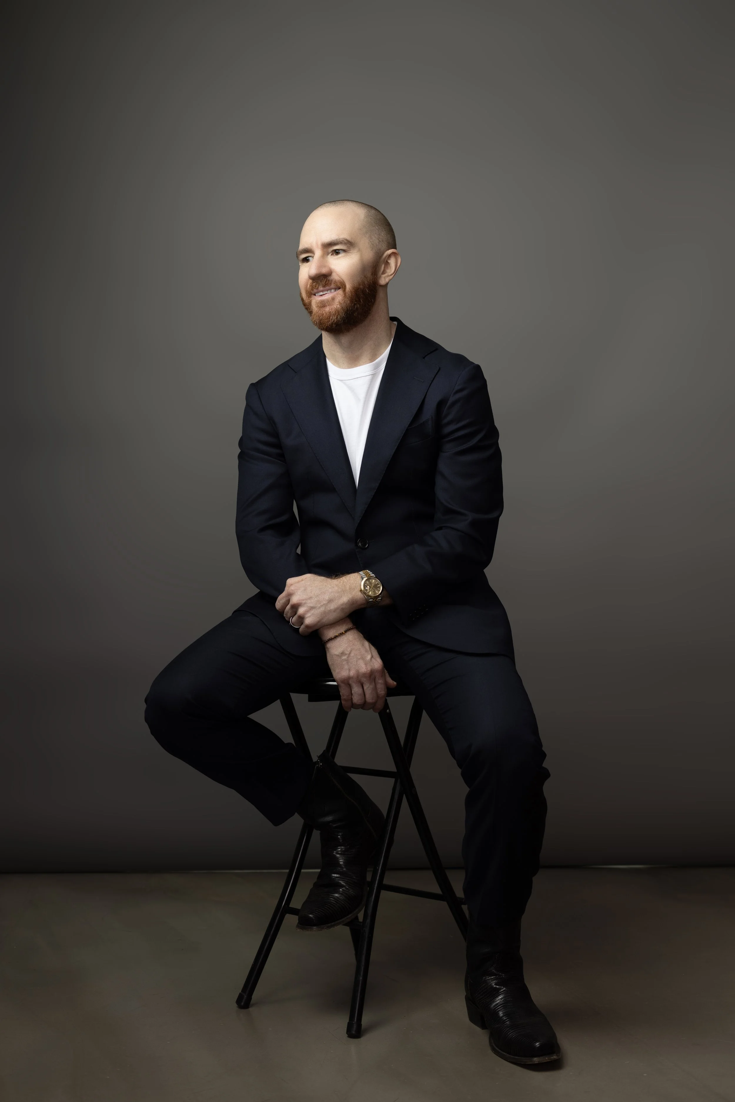 A man with a beard and a shaved head, wearing a dark blazer, white t-shirt, and black pants, seated on a stool against a grey background, smiling slightly and looking to his right.