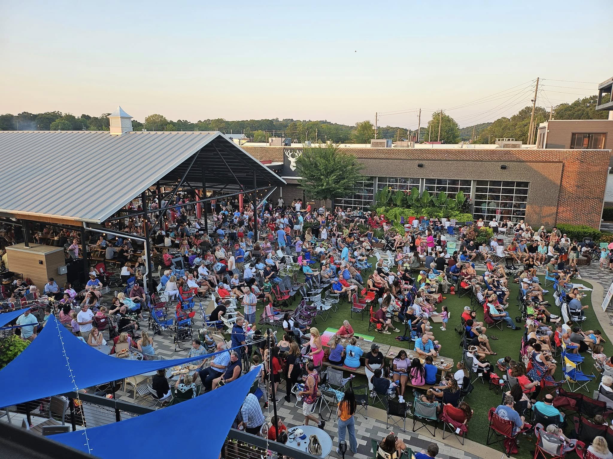 A large outdoor gathering with many people seated on chairs and at tables in a courtyard. There is a covered pavilion on the left side and a building with large windowpanes and greenery on the right. The crowd appears to be enjoying an event or conce