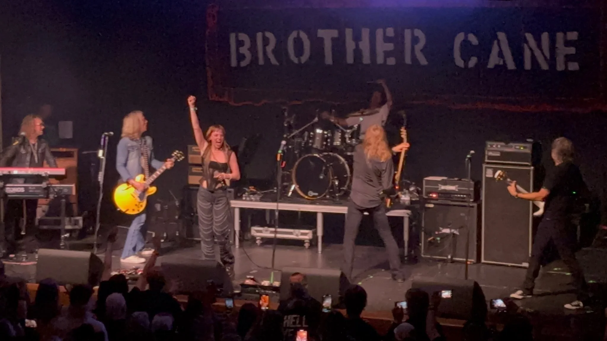 A band performing on stage with six members, one with a keyboard, one with a yellow electric guitar, a female singer holding a microphone, a bassist with an orange bass guitar, a drummer in the background, and a guitarist on the right. Audience membe