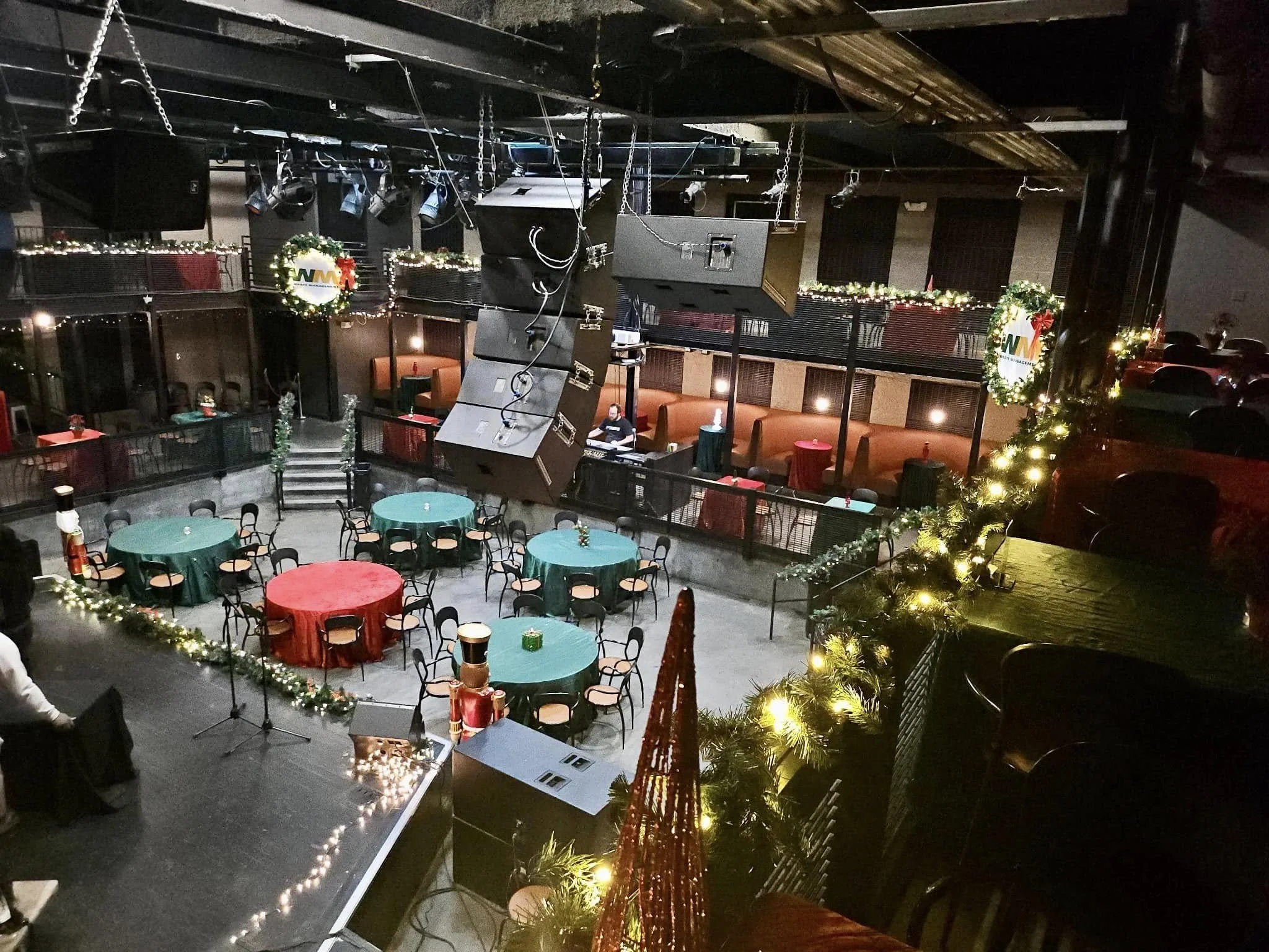 The image shows a decorated indoor event venue with round tables covered in red and green tablecloths, surrounded by black chairs. The room features Christmas decorations, garlands, and wreaths with lights. There are large hanging speakers and a smal