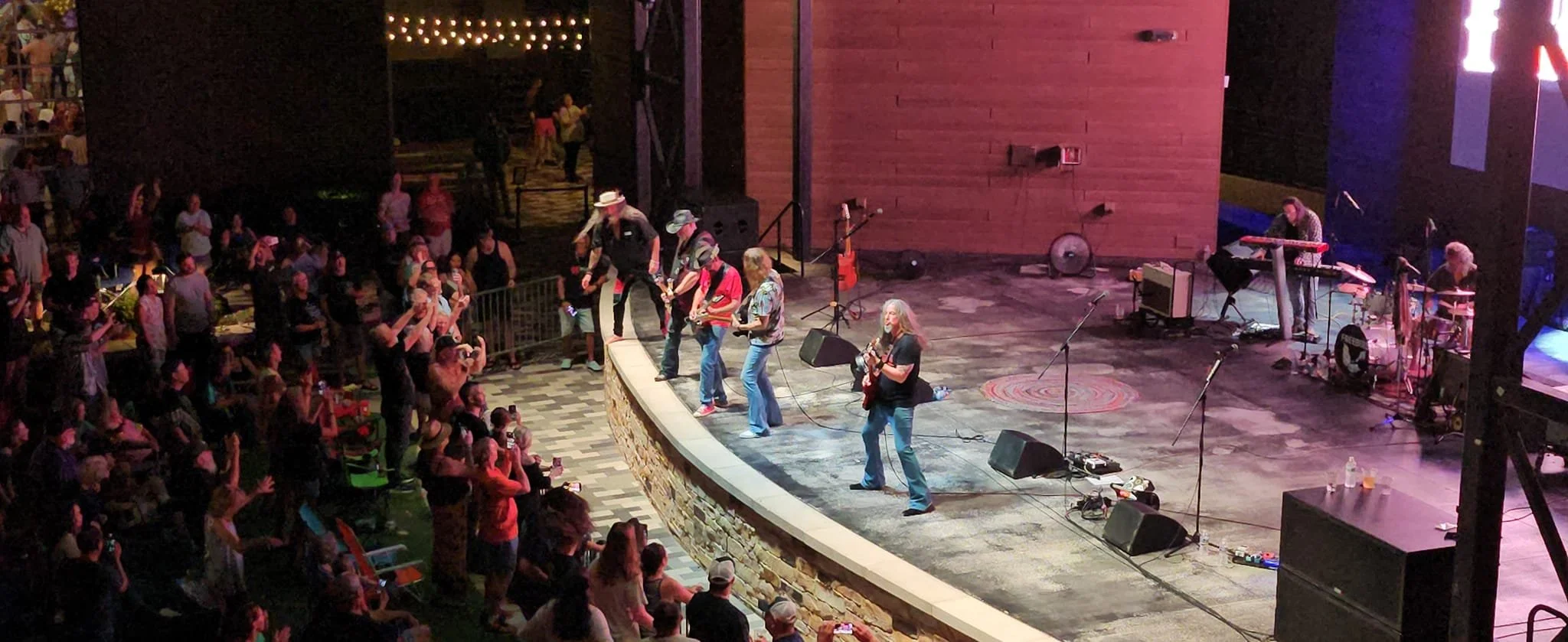 A live outdoor music concert with a band performing on stage and an audience in front of them. The band includes guitarists and a keyboard player. Audience members are watching and taking photos, with some standing and others seated.