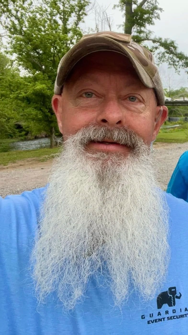 A man with a long white beard and a camouflage baseball cap taking a selfie outdoors, with trees and a river in the background.