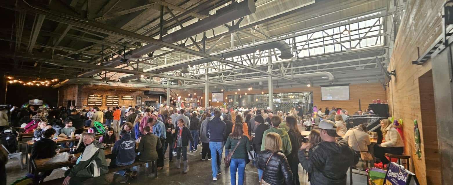 Crowd of people inside a large industrial-style bar or event space with high ceilings, metal beams, and wooden walls, some sitting and some standing, with a bar area in the background.