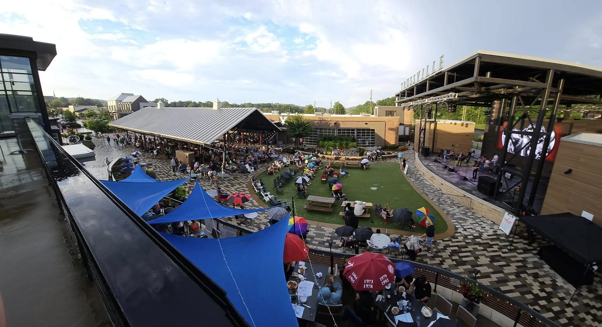 An outdoor concert at a shopping or entertainment complex with a large stage on the right and an audience with umbrellas and seating throughout the area. There are tents and buildings in the background.
