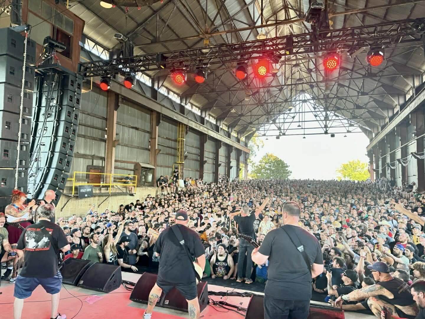 A band performing on stage at an indoor concert with a large audience in front, under a high metal roof with red stage lights.