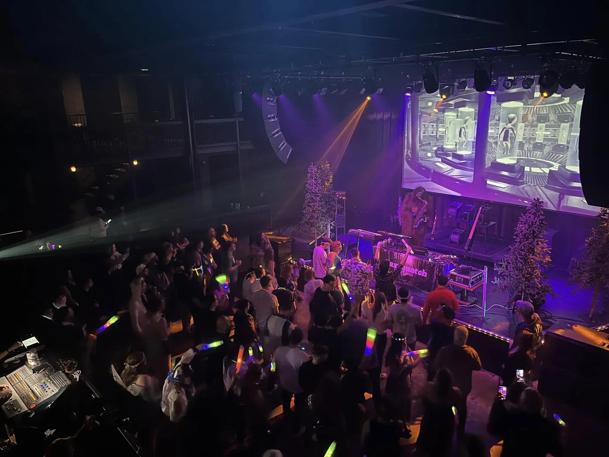 View of a nightclub with a crowd dancing on the floor in front of a DJ booth. The DJ is playing electronic music, and the stage features large digital screens with futuristic visuals. Colorful lights and winter-themed trees decorated with snow add to