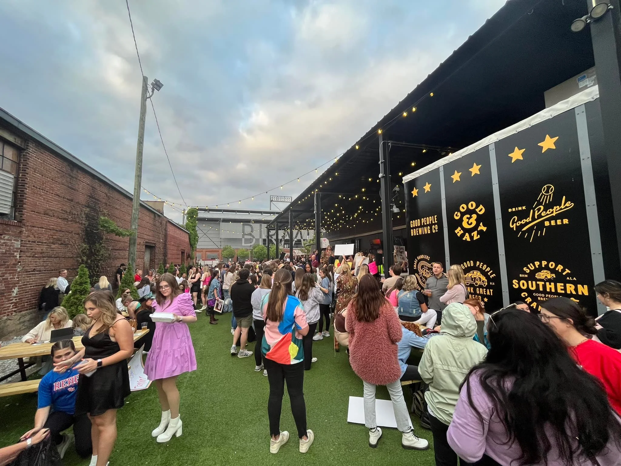 Crowd of people gathered outside a brewery with string lights overhead, some sitting and some standing, under a black awning that reads 'Good People Brewing Co.' and 'Drink Good Beer' with yellow star graphics.