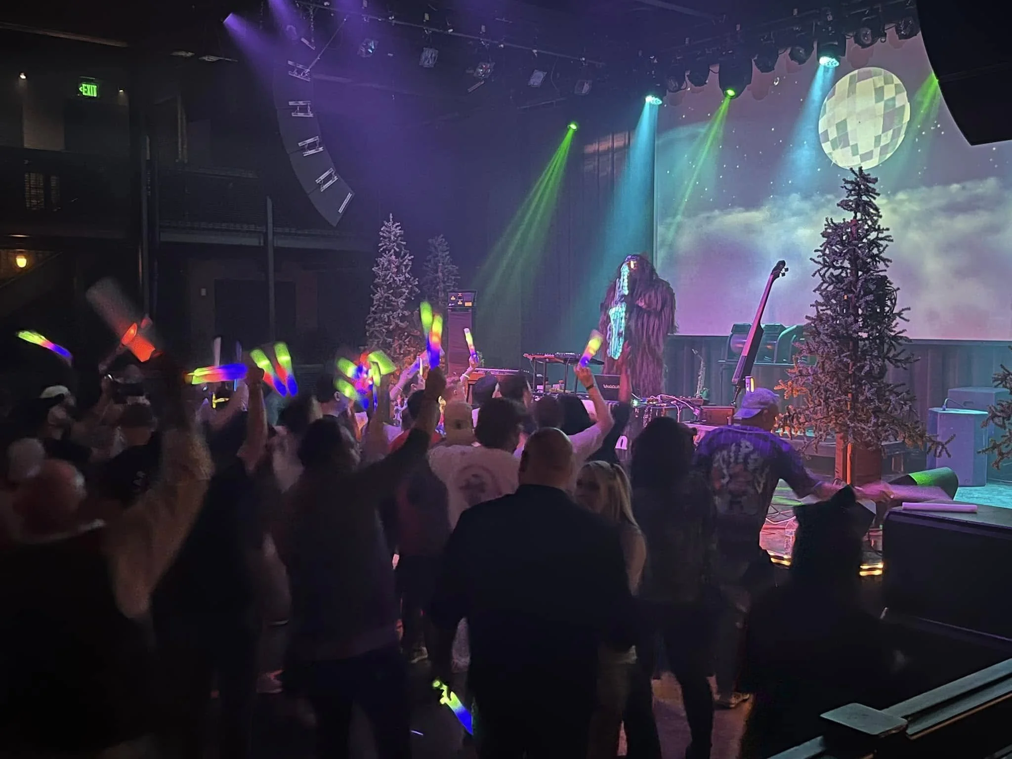 A lively concert or party scene with a DJ or performer on stage, colorful lights, and a crowd dancing with glow sticks. The background has Christmas trees and a large projection of the moon in a night sky with clouds.