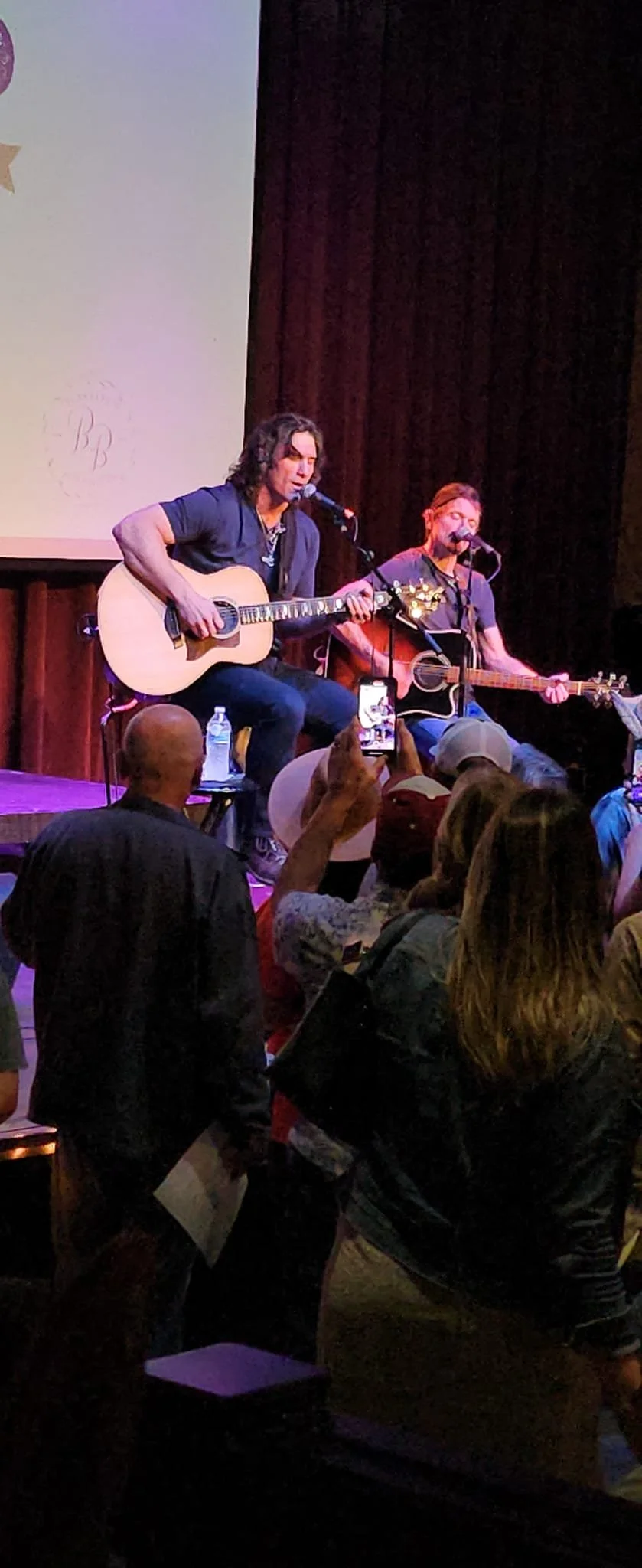 Two musicians are playing acoustic guitars and singing on stage during a performance. The stage has a dark curtain backdrop, and there is a large audience in front.