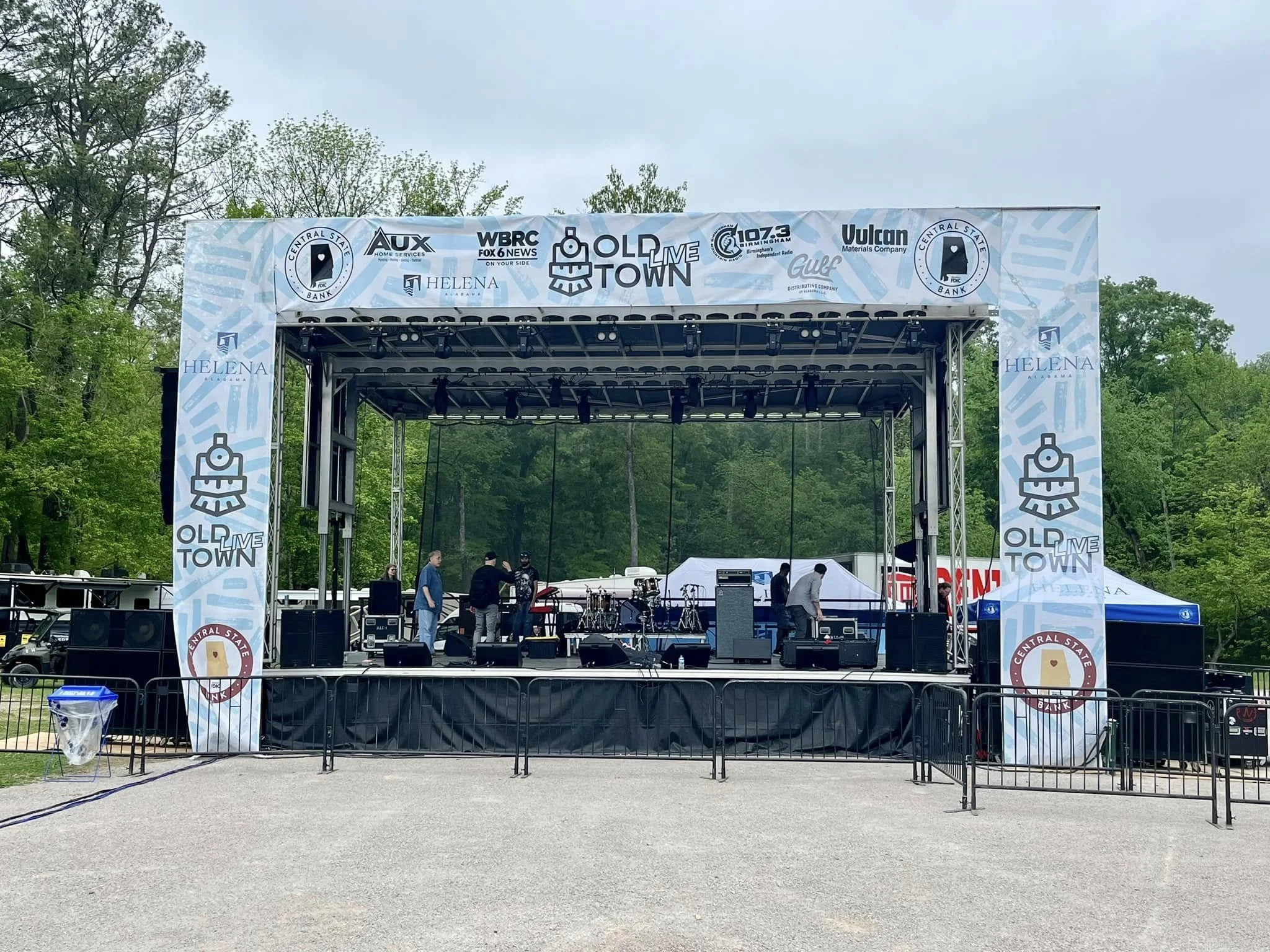Empty outdoor stage set up for a live event, with sound equipment and crew preparing, surrounded by green trees and overcast sky.