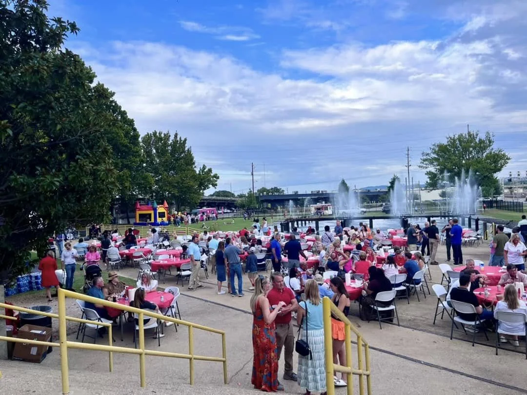 People gathered at an outdoor event with pink tables, a fountain, and bounce houses in the background under a partly cloudy sky.