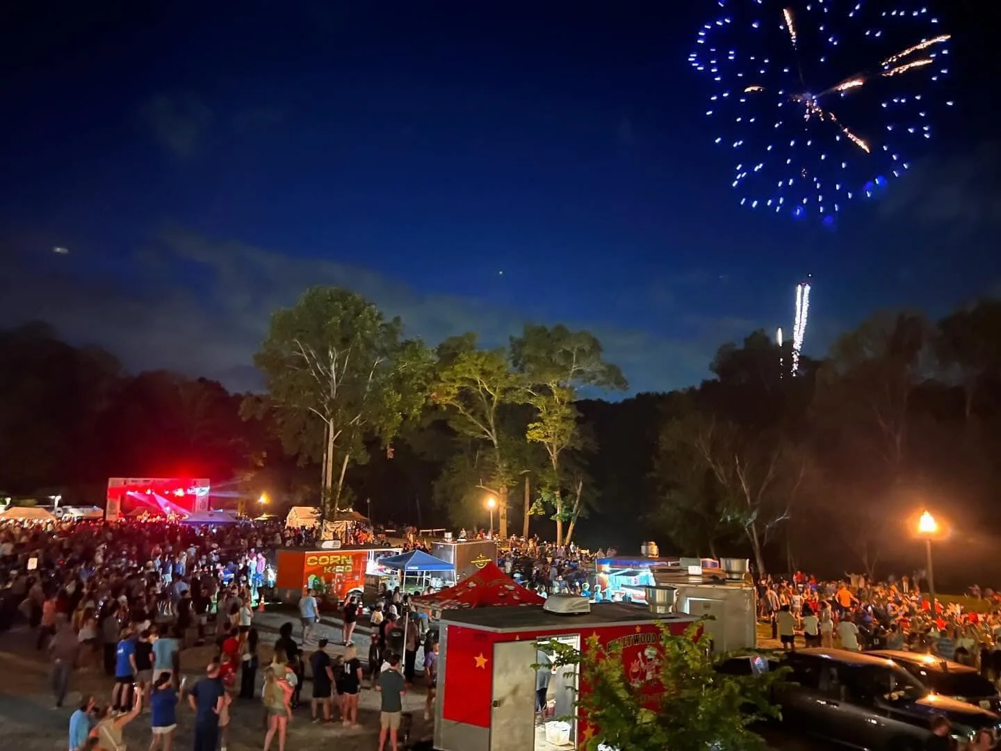 Nighttime outdoor fair with a large crowd, food stalls, and a stage with a red light. Fireworks display in the sky above trees.