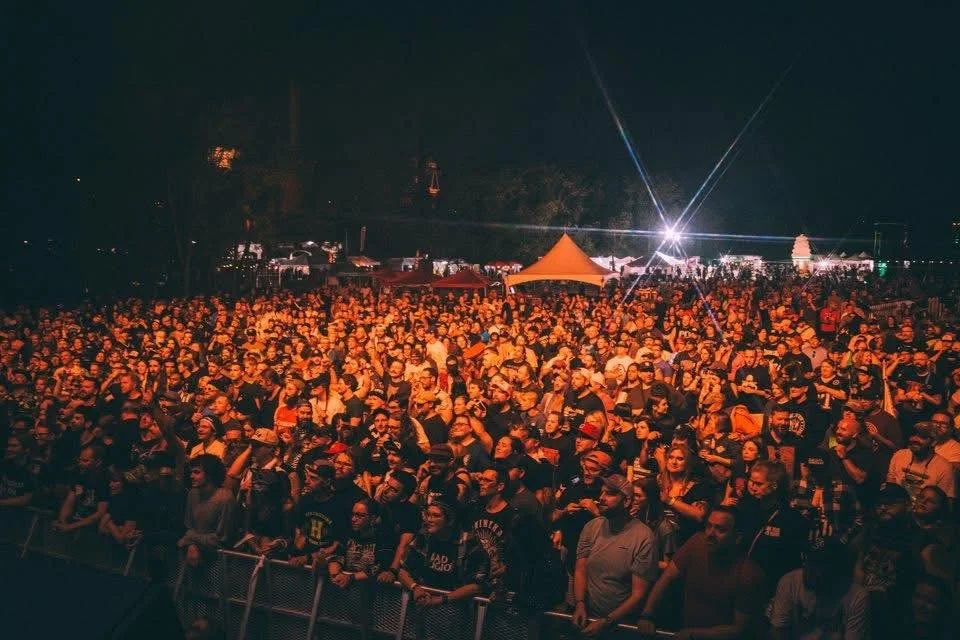 Large crowd gathered at an outdoor night event or concert with stage lighting and tents in the background.