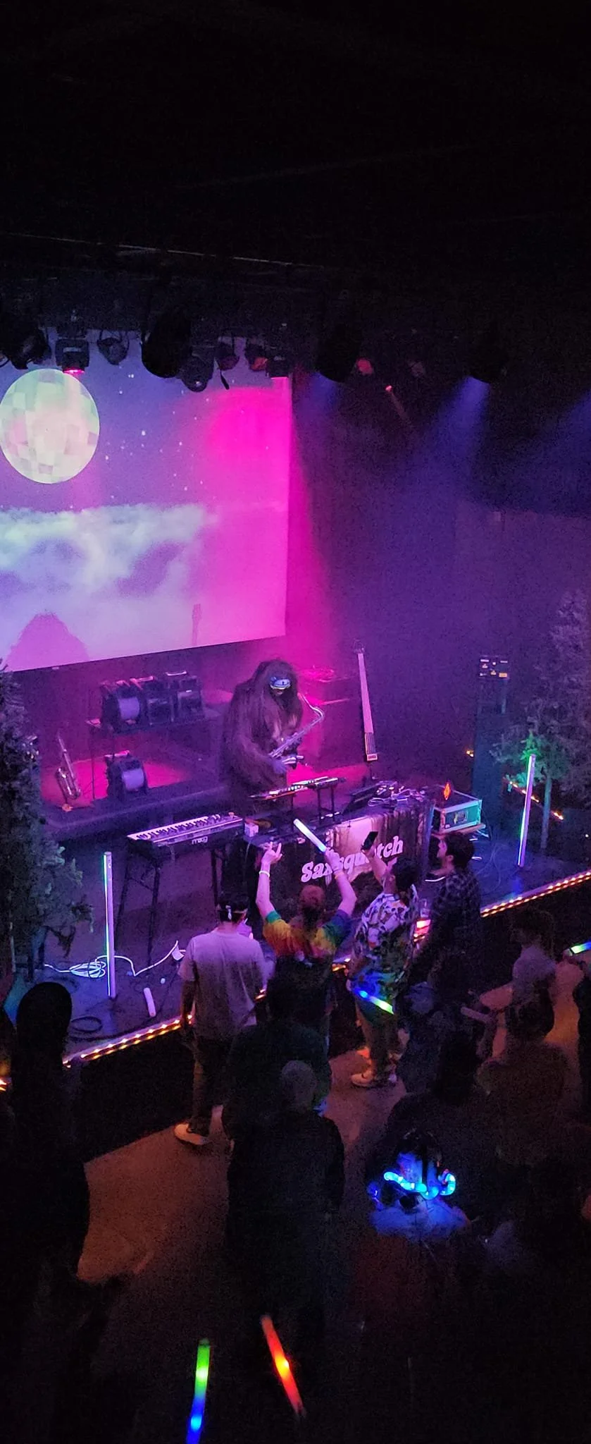 People dancing and enjoying a nightclub or concert with a DJ on stage, colorful lighting, neon glow sticks, and a large screen displaying a moon and clouds.