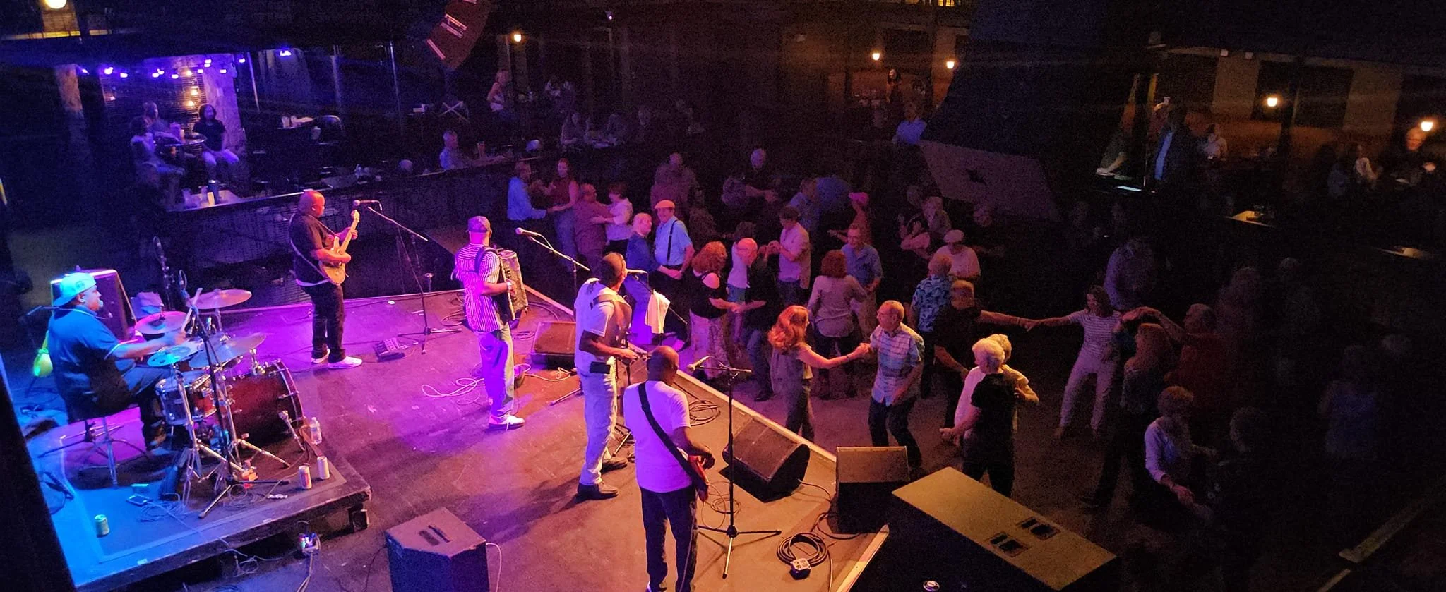 A live band performs on stage at a social dance event where people are dancing in pairs on a dimly lit dance floor.