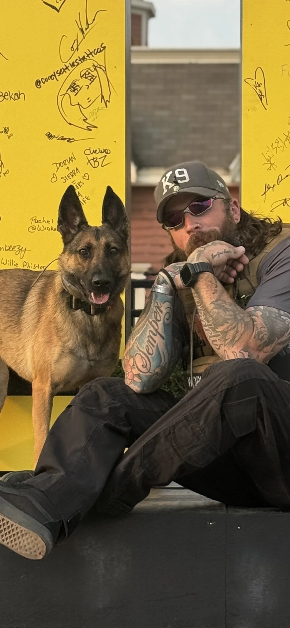 A man with long hair, sunglasses, and tattoos sitting on the ground next to a Belgian Malinois dog, both positioned in front of a yellow wall with handwritten signatures and drawings.