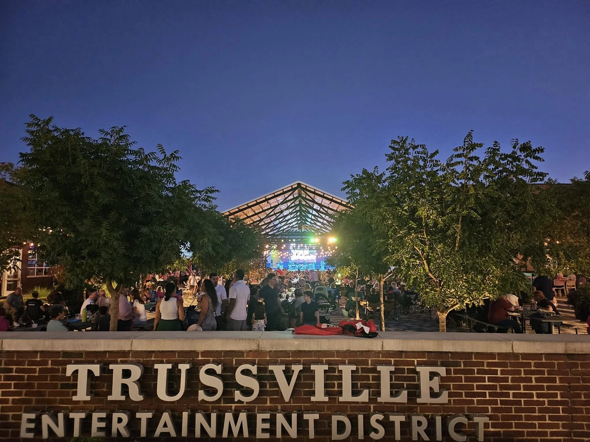 A lively outdoor scene at Trussville Entertainment District with a stage and colorful lights, surrounded by trees and people socializing during the evening.