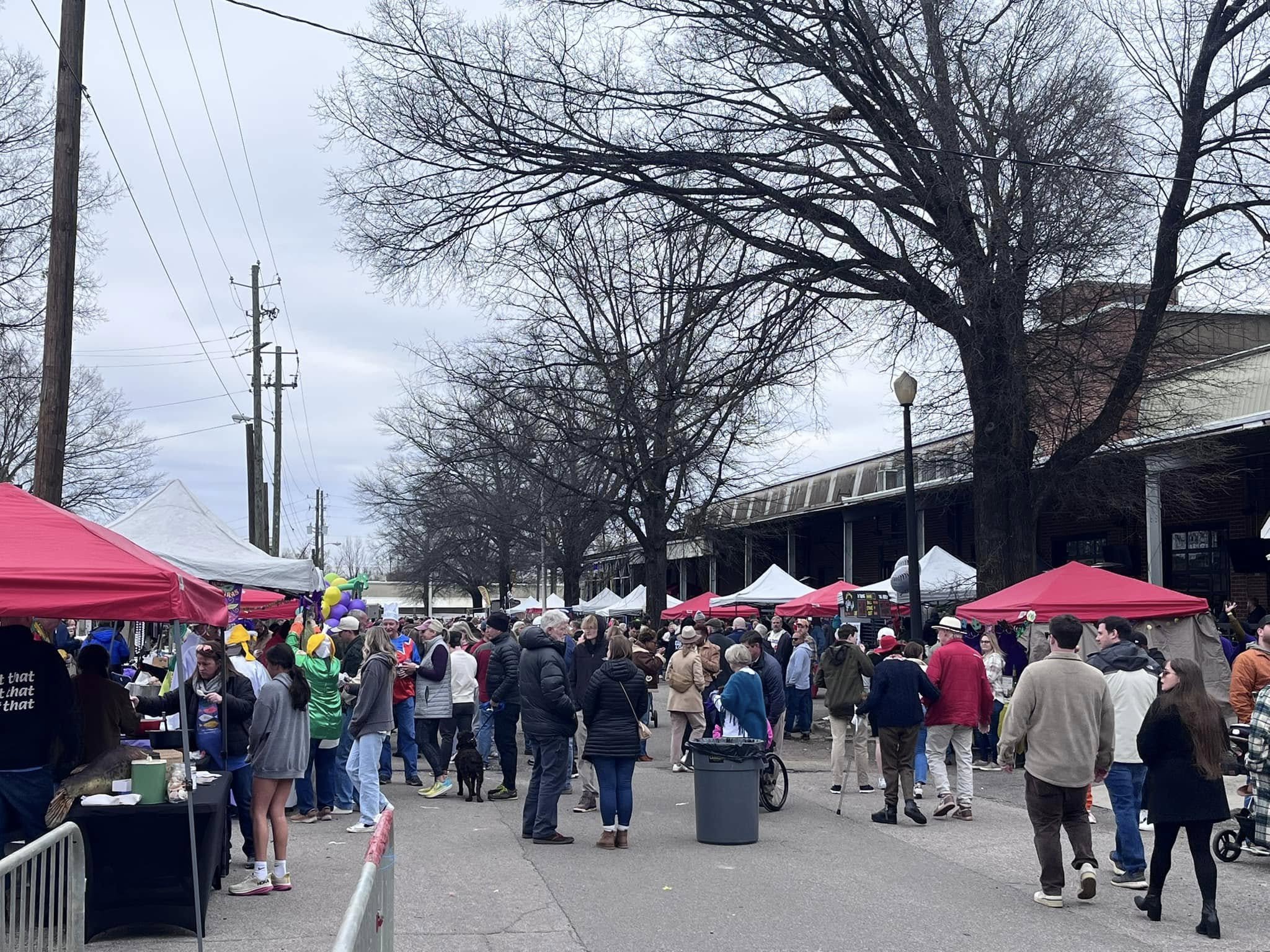 A street fair with many people walking among vendor tents. The tents are white and red, and some are decorated with balloons. The weather is overcast, and leafless trees line the street.