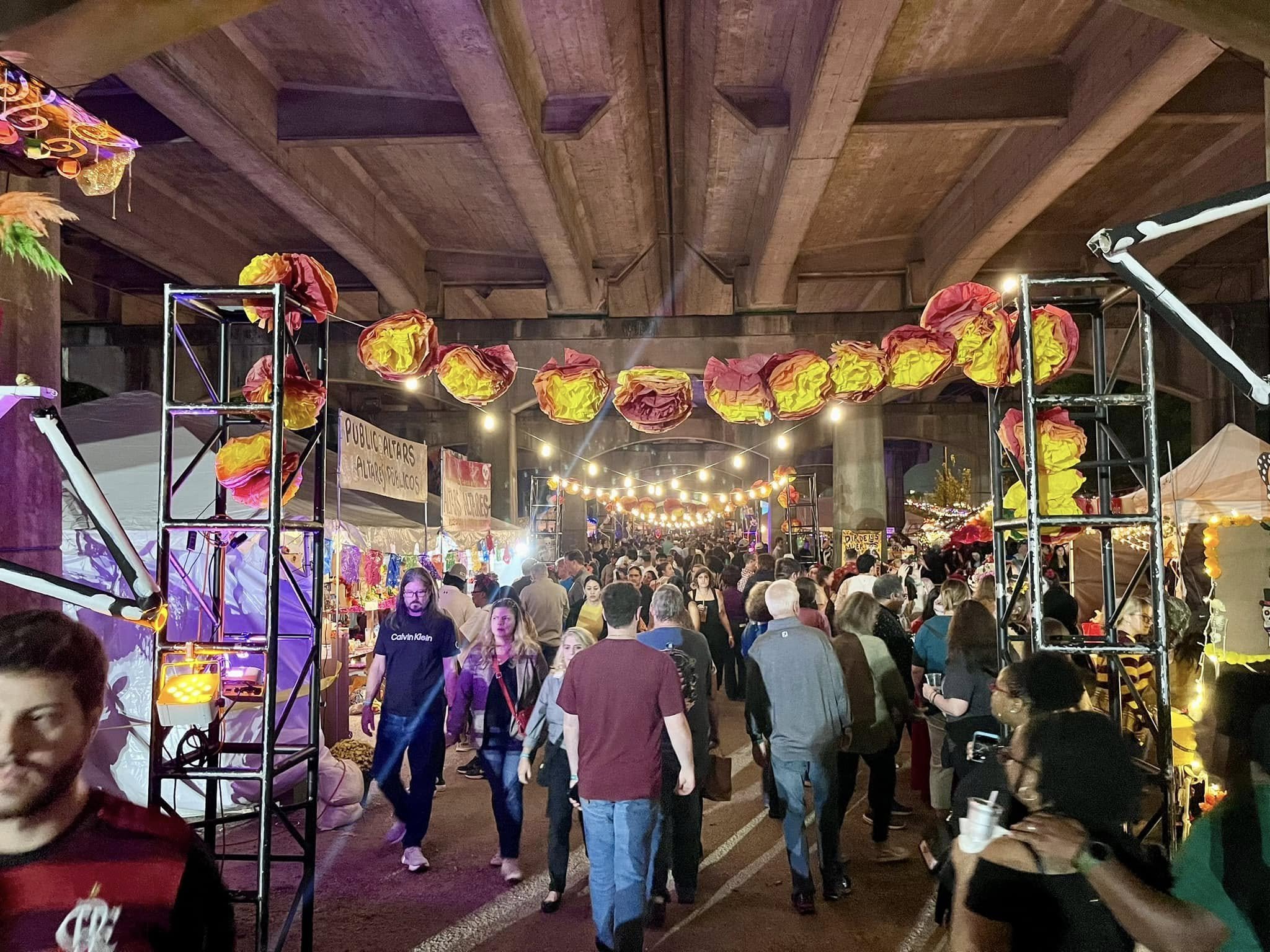 Crowded street fair or festival under a bridge, decorated with illuminated paper lanterns and string lights, with various vendor stalls and multiple people walking and browsing.