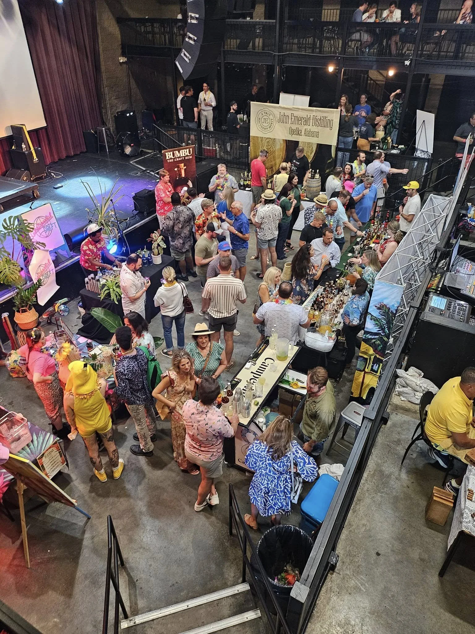 An indoor event showing a crowded market or festival with a stage on the left and various activity booths. People are walking around, talking, and drinking. There are banners, including one for John Emerald Distilling in Opelika, Alabama. The scene i