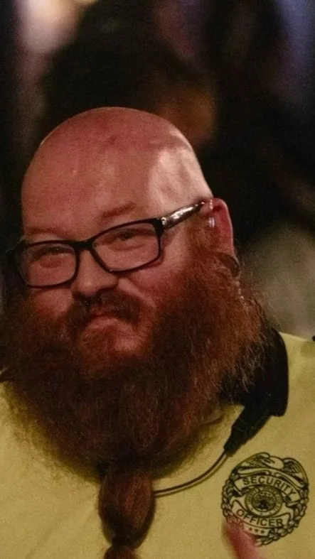 A man with a bald head, glasses, and a long red beard styled in a braid. He is wearing a security officer shirt and is in a dimly lit environment.