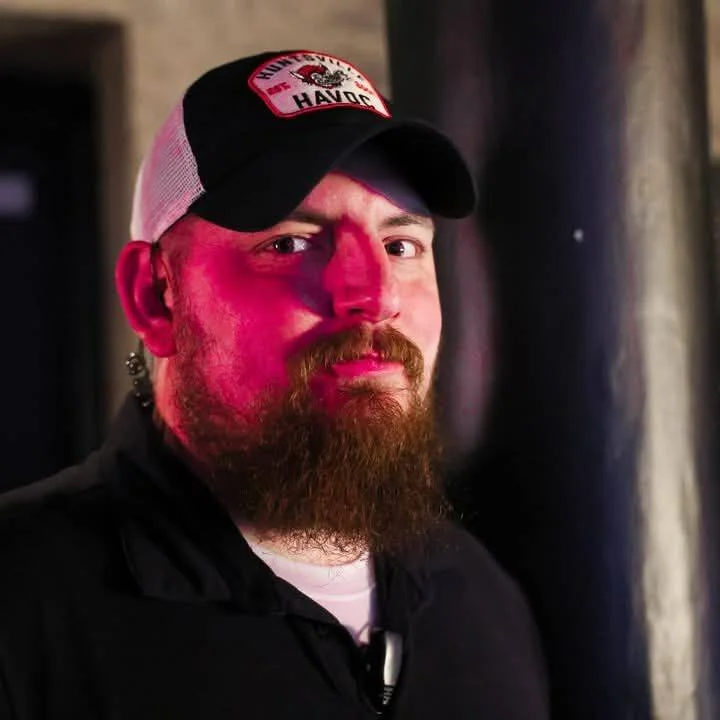 A man with a red beard wearing a black and white trucker hat with a patch that says 'HARD CORE HAVOC'.