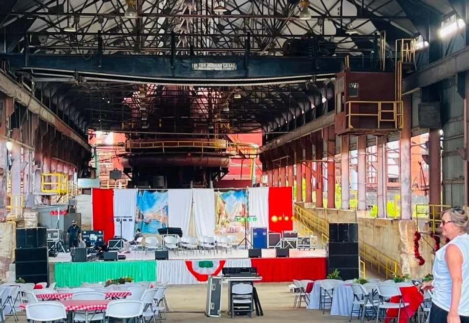 An indoor event space set up with decorated tables and chairs in front of a stage, with a large industrial building structure overhead.