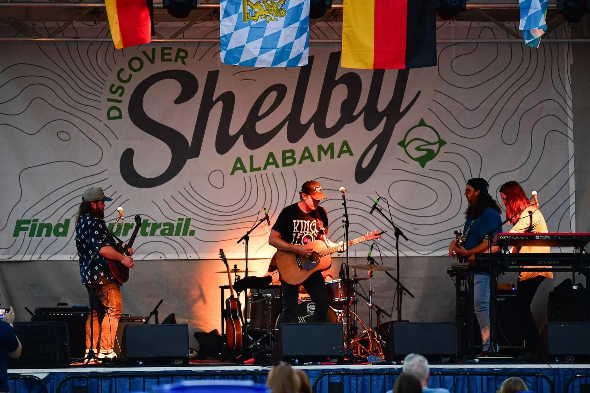 A band performing on stage at an outdoor event called 'Discoer Shelby Alabama,' with three musicians playing guitar and keyboard. The background features a large banner with the event name and flags hanging above.