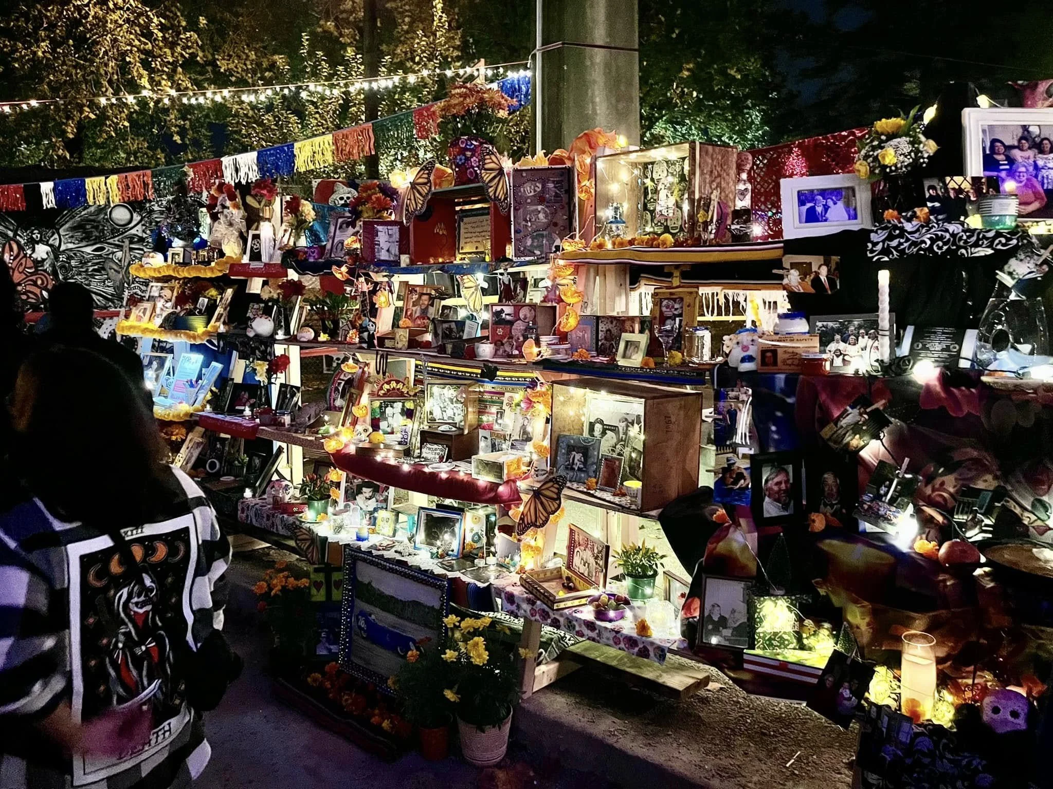 Nighttime ofrenda decorated with photos, candles, flowers, and candles, with strings of paper flags above and trees in the background.