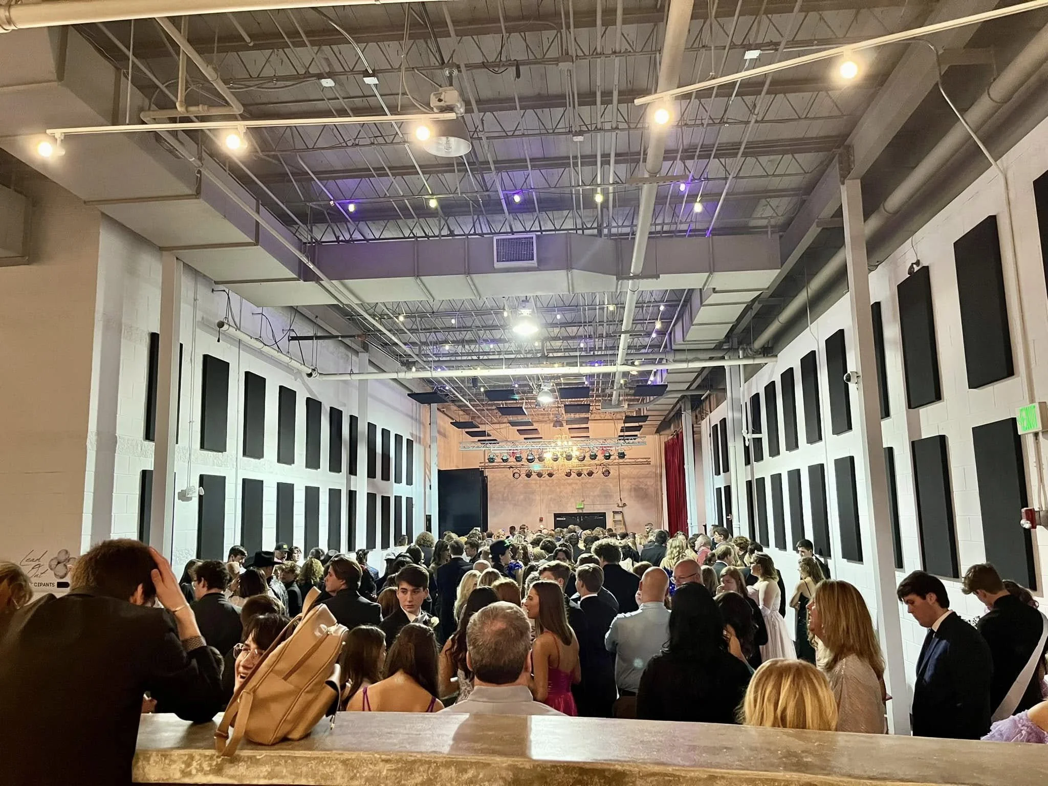 Crowd of people gathers in a large indoor space with a stage at the front, under a high ceiling with exposed pipes and lighting.