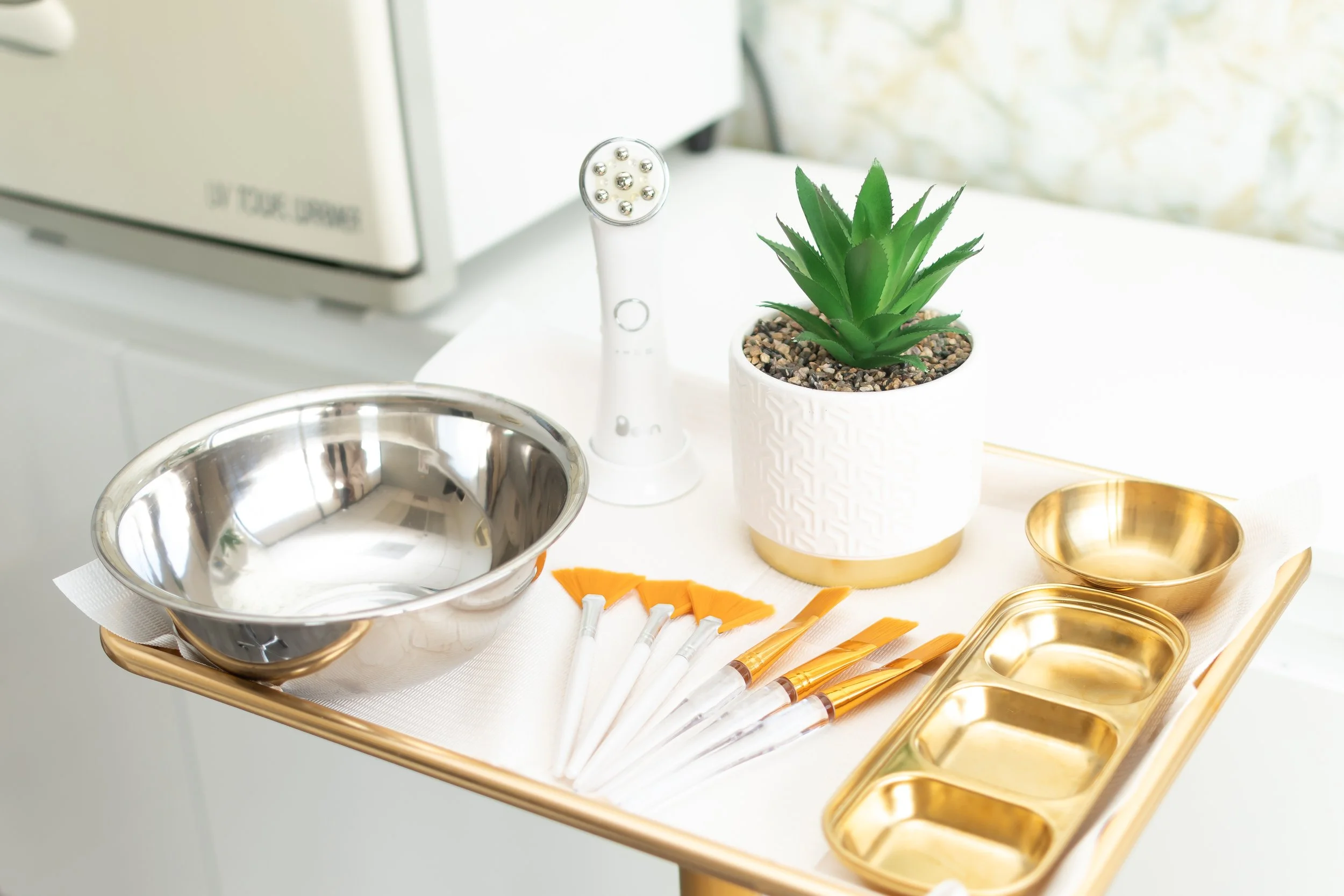 A tray with a potted green succulent plant, a set of paintbrushes with orange and gold handles, a metallic mixing bowl, a set of small gold bowls, a gold divided dish, a white tool with multiple ball bearings, and a white cloth, placed next to a microwave oven.