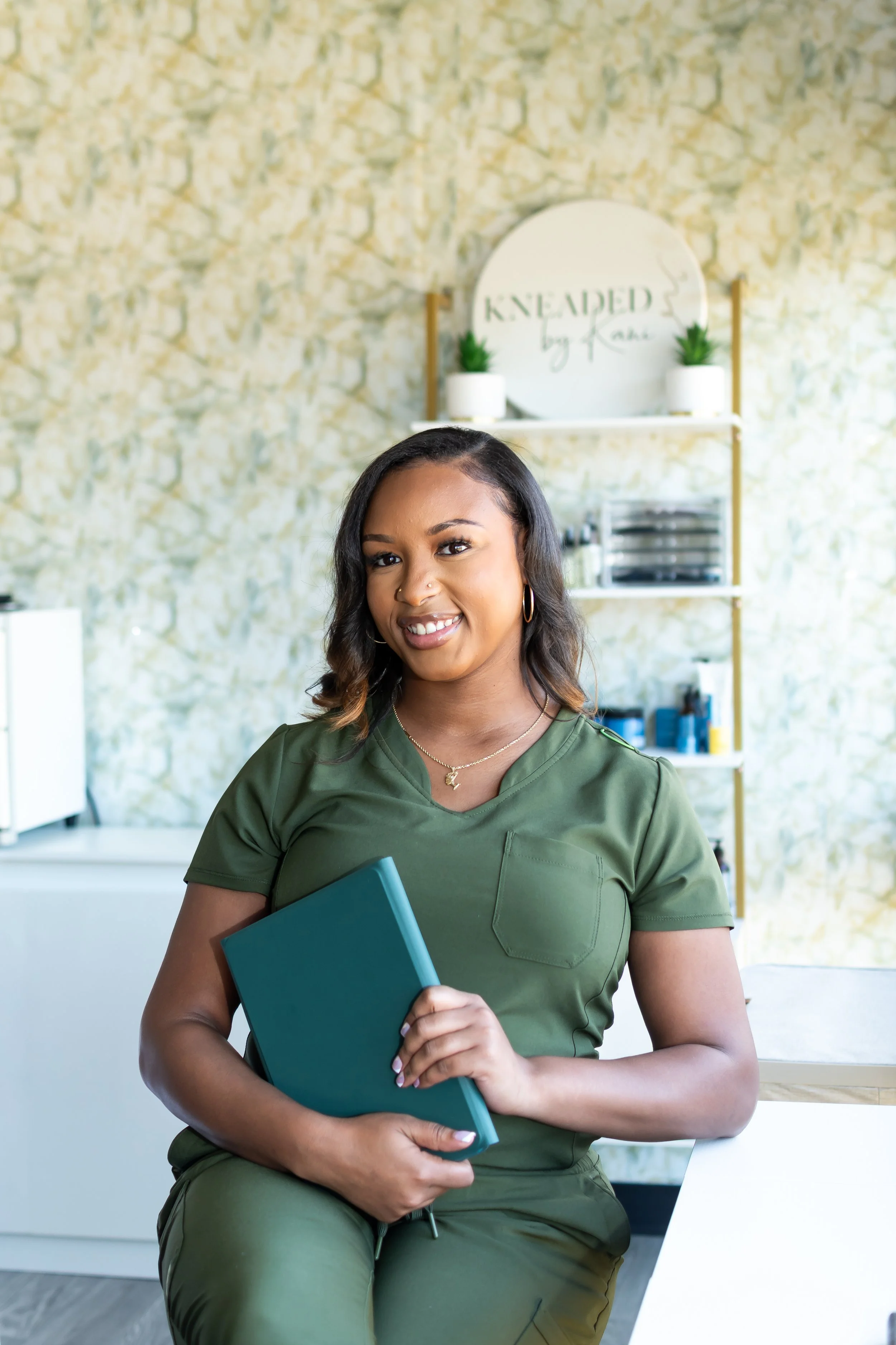 A smiling woman in green scrubs sitting in a nail salon, holding a teal folder with shelves and a sign that reads "Kneaded by Rae" in the background.