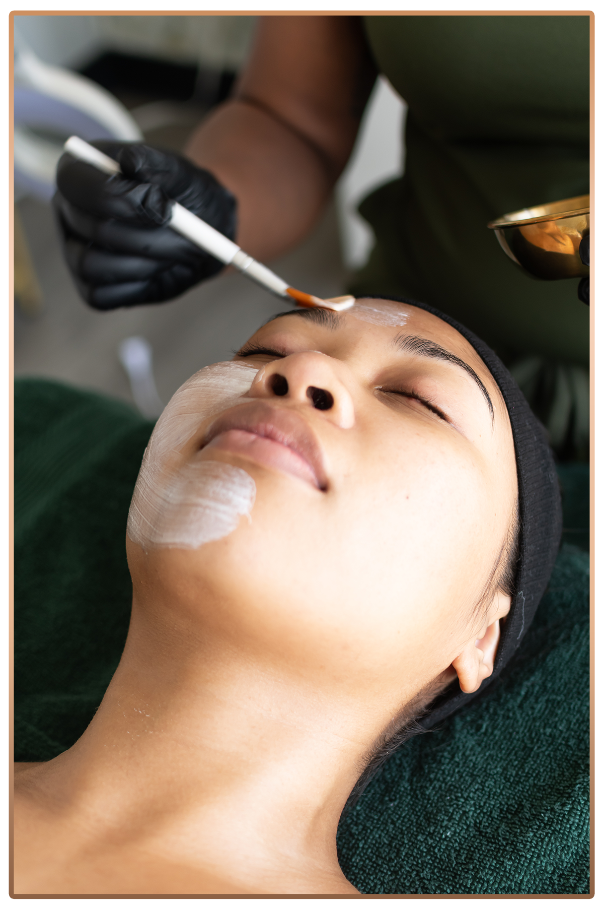 A woman receiving a facial treatment, lying down with her eyes closed, while a skincare professional applies a mask to her forehead using a brush.