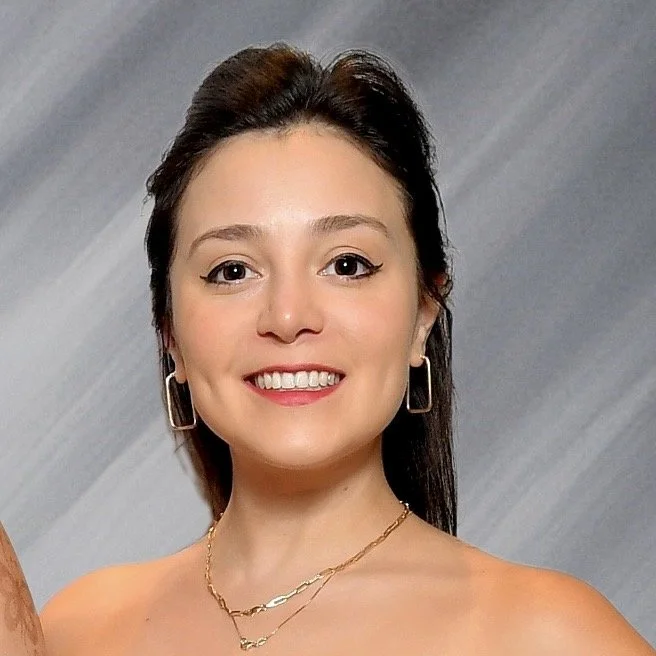 Close-up portrait of a smiling woman with dark hair, hoop earrings, a gold necklace, and a neutral background.
