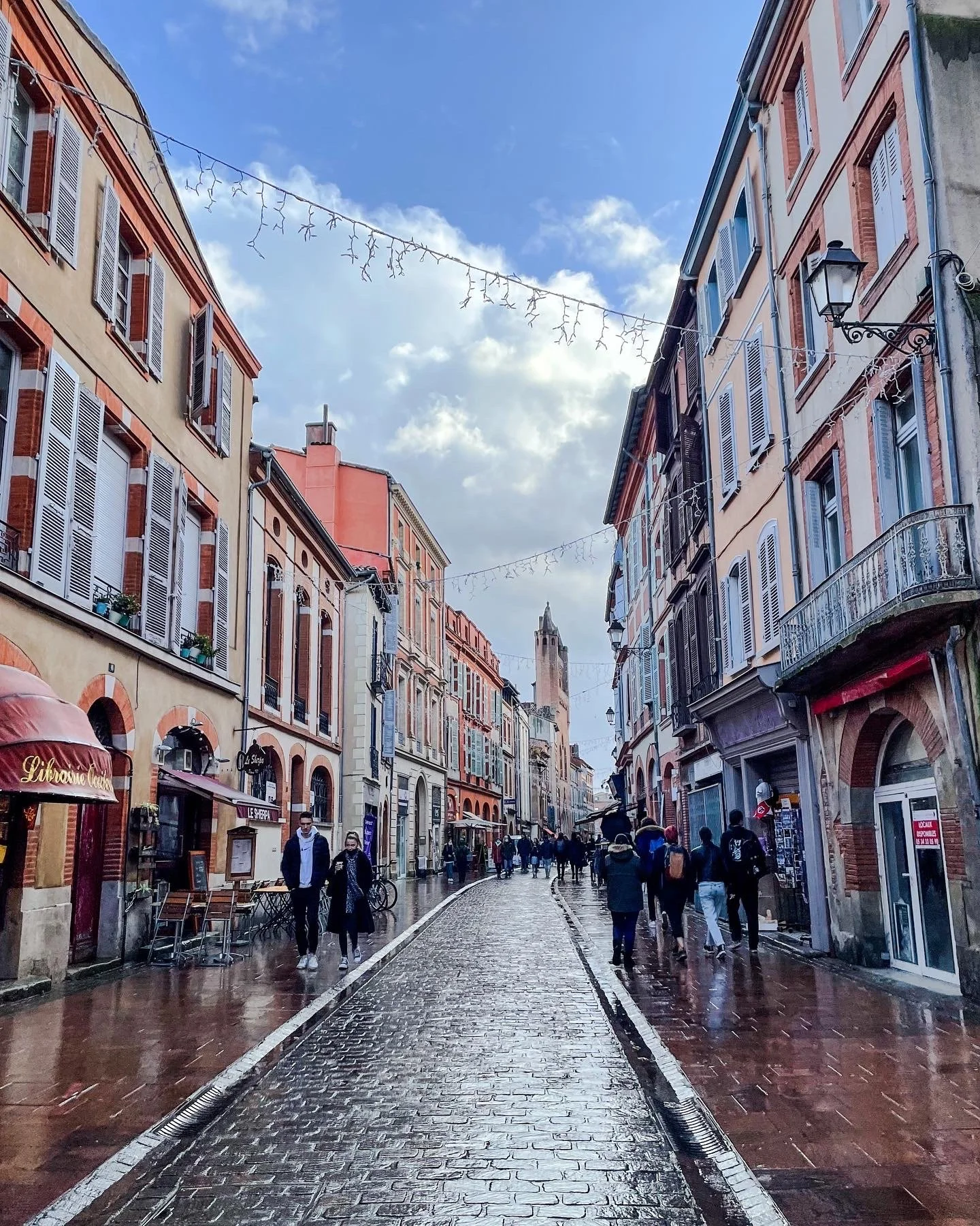 A cobblestone street lined with colorful buildings, some with shutters, under a partly cloudy sky. Pedestrians walk along the street, and there are cafes and shops on either side. String lights hang above the street.