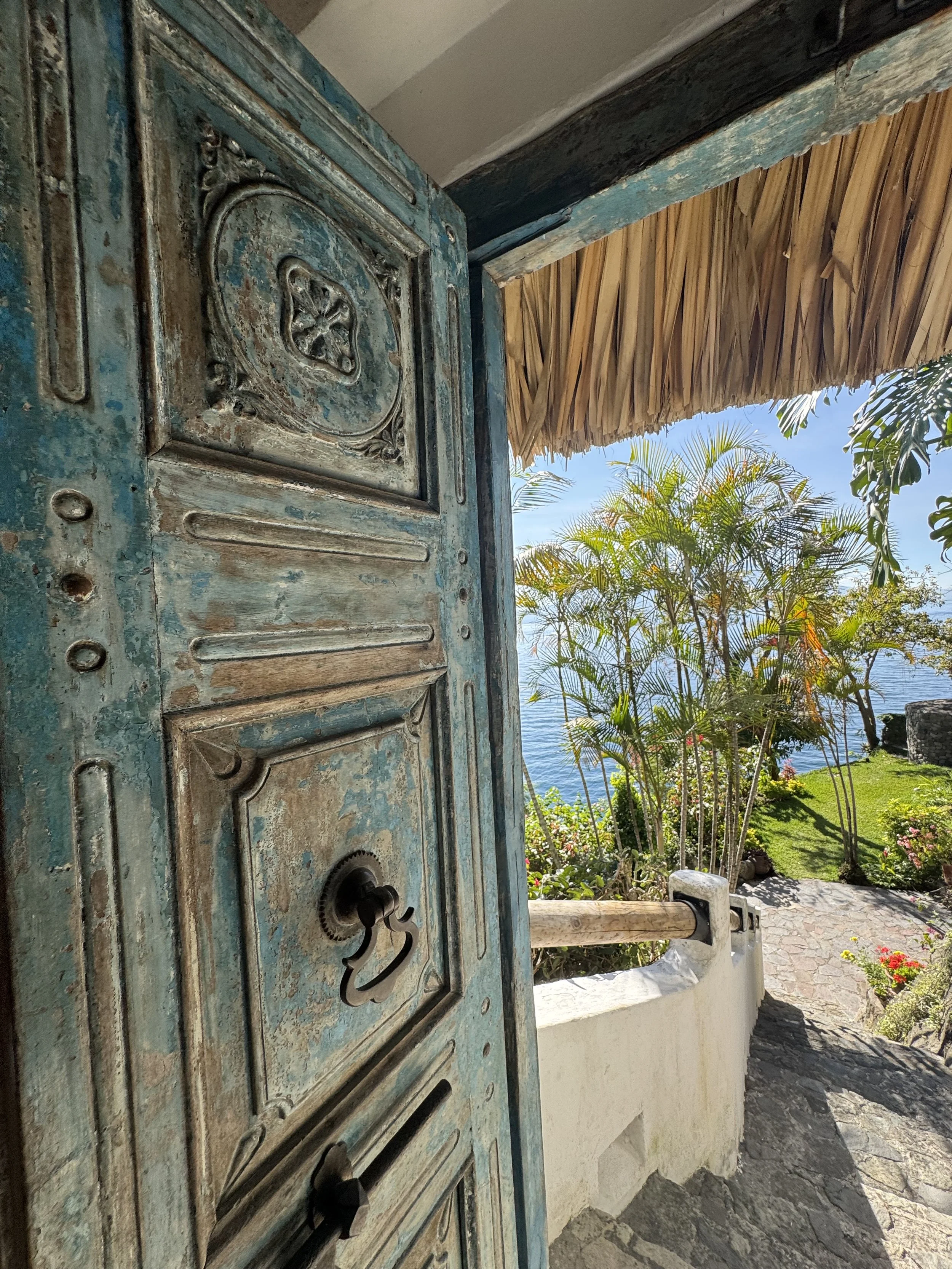 Open rustic turquoise door with ornate wood paneling, leading to a tropical garden patio overlooking the ocean with palm trees and flowers.