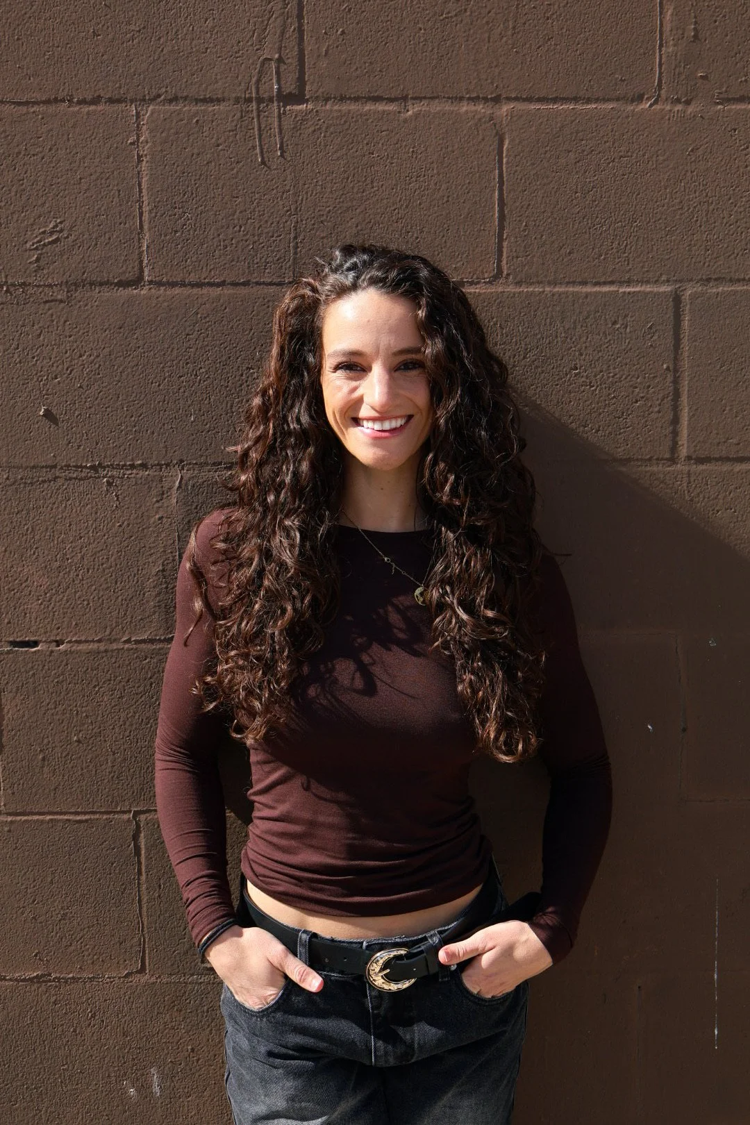 A woman with long curly dark hair smiling, standing against a brick wall, wearing a long-sleeve brown top and dark pants with a belt.