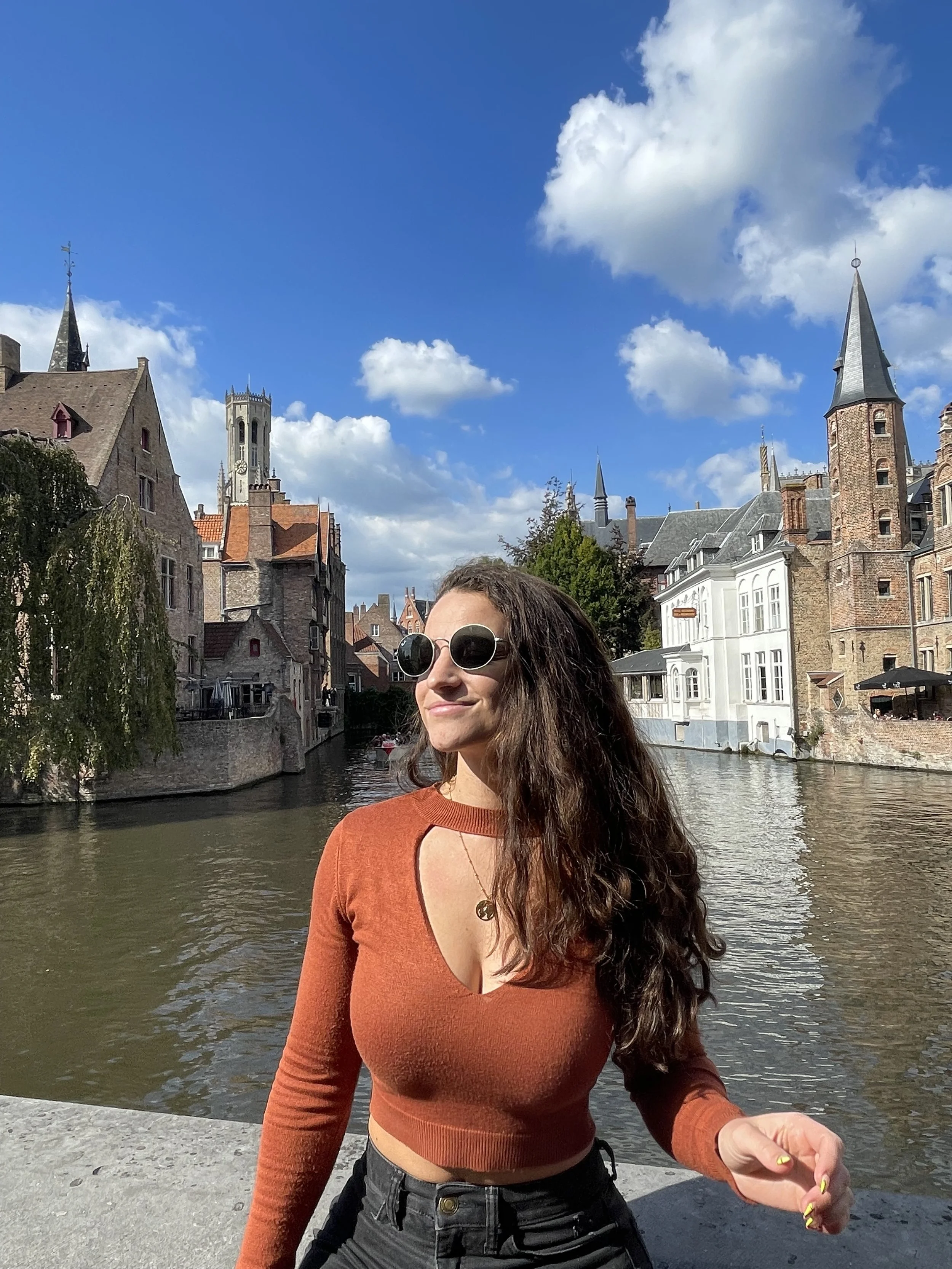 A woman with long, curly brown hair wearing sunglasses, a rust-colored crop top with a keyhole cutout, and black pants, standing by a canal in a historic European city with medieval buildings and towers under a partly cloudy sky.