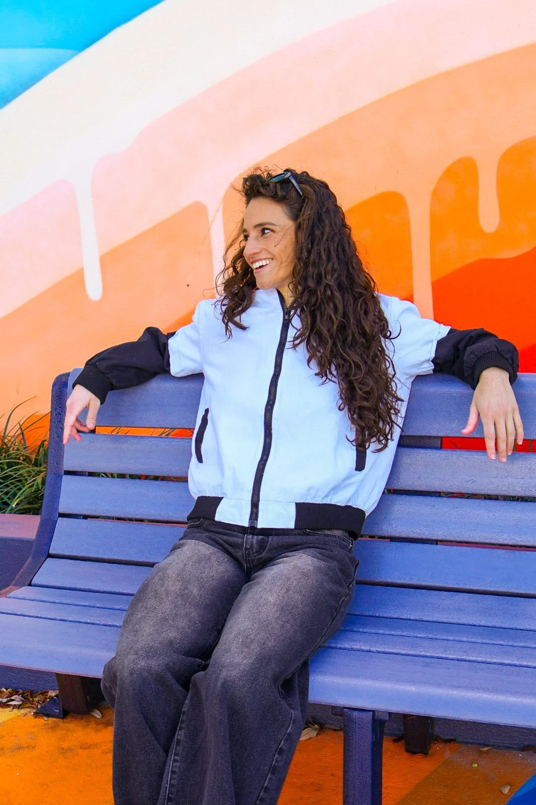 A woman with long, curly hair sitting on a blue park bench against a colorful mural background.