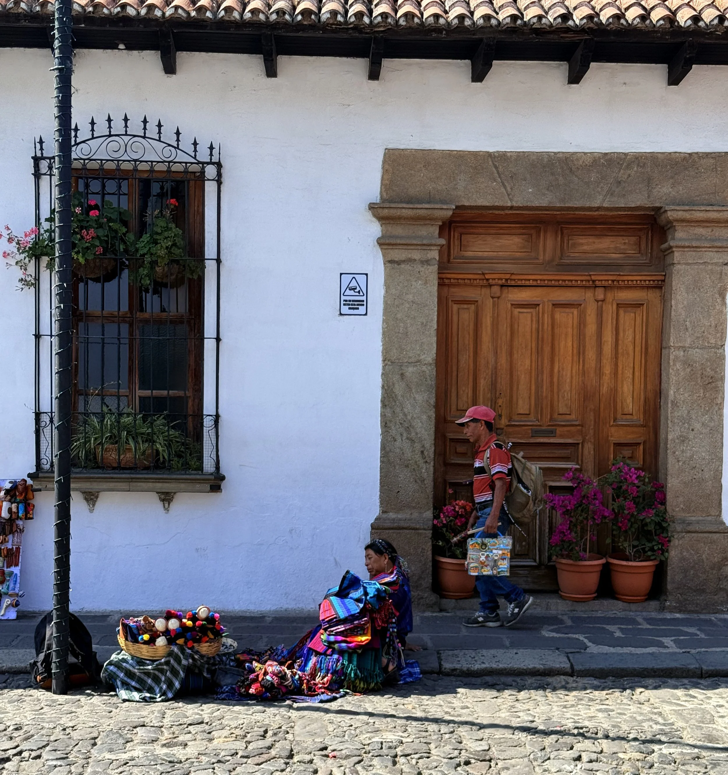 Street scene with a woman selling colorful textiles and a man walking past, both wearing traditional clothing, in front of a white building with a wooden door and potted flowers.