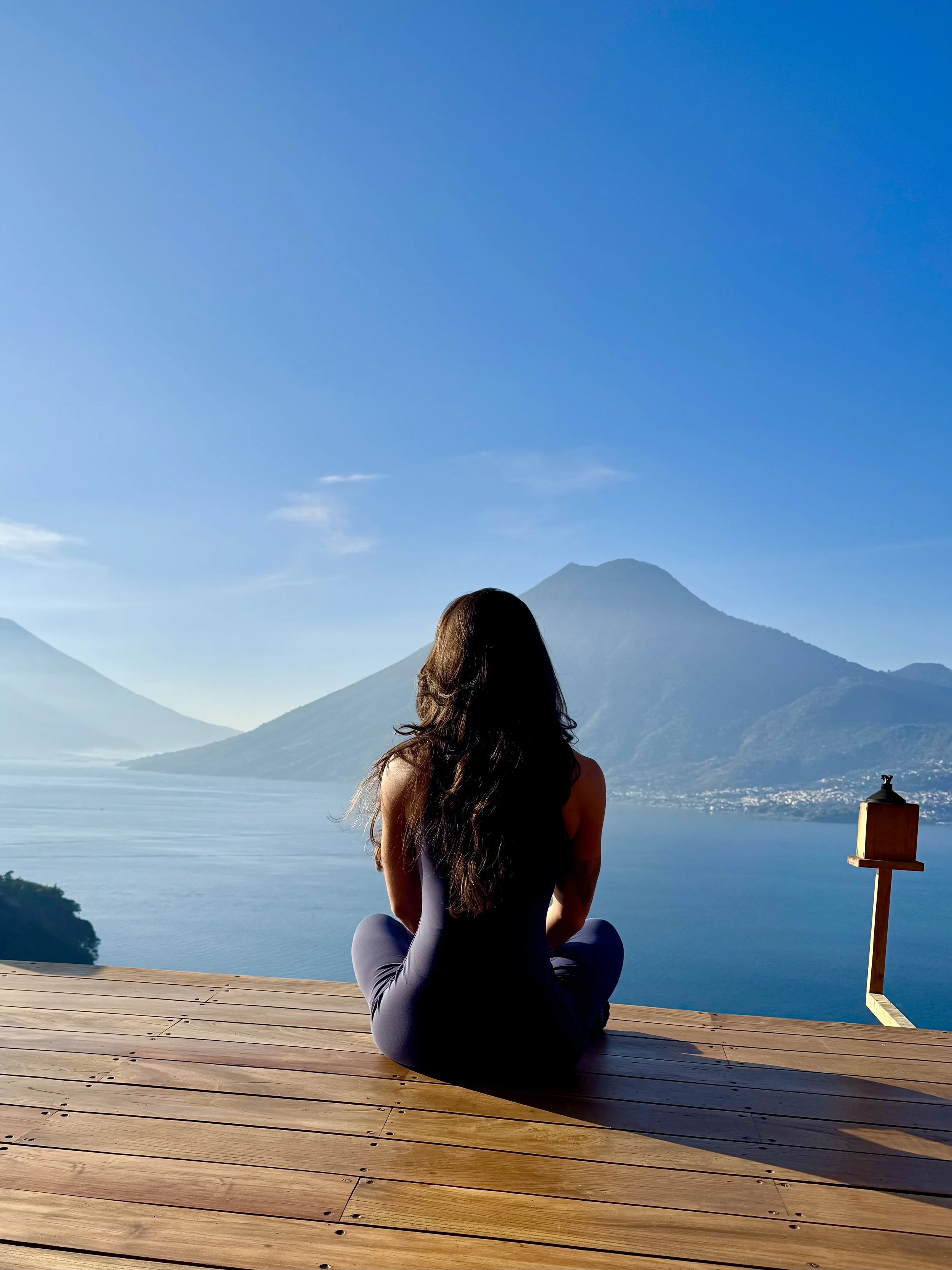 A woman with long dark hair sitting cross-legged on a wooden deck, facing a mountain and lake landscape under a clear blue sky.