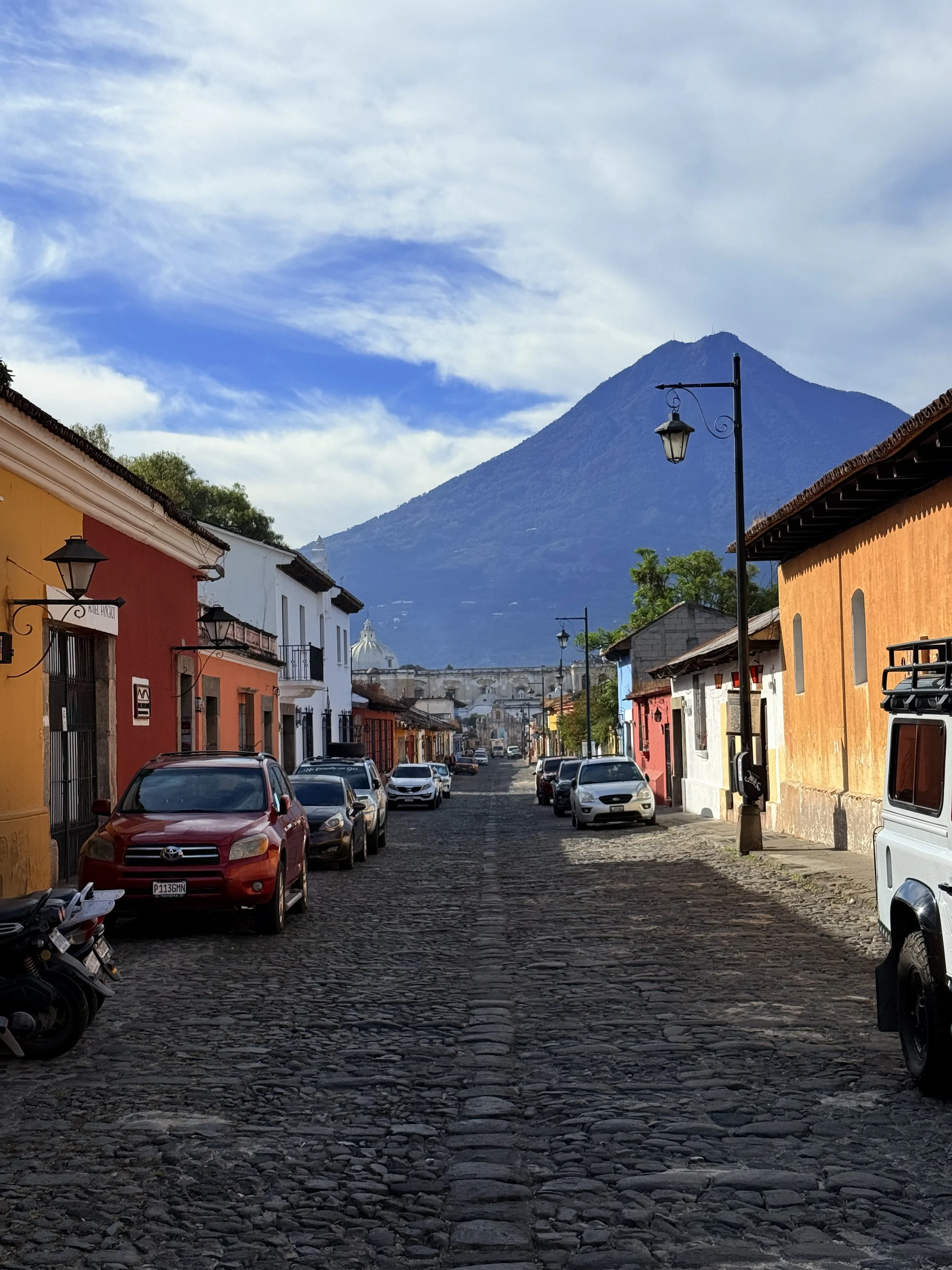 A cobblestone street in a colorful town with parked cars and buildings, with a large mountain in the background and a partly cloudy sky.