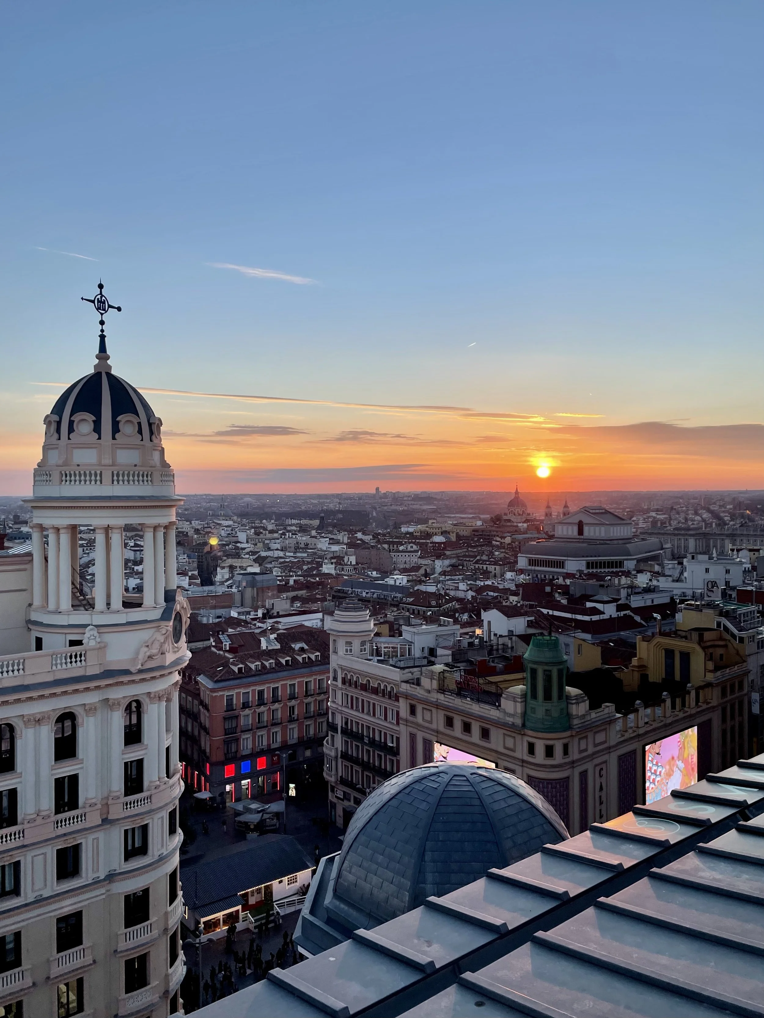 Cityscape view at sunset with historic buildings and a dome, showcasing a skyline with the sun setting near the horizon.