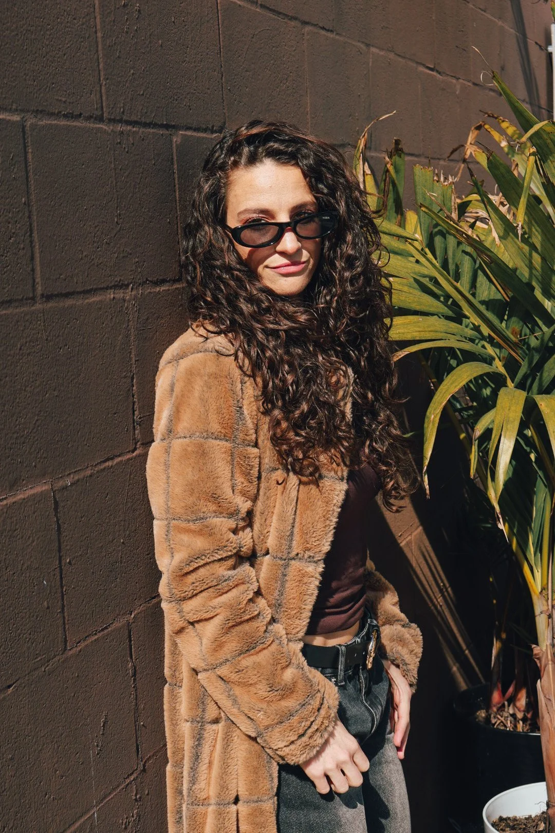 Young woman with curly hair wearing sunglasses and a brown fur coat standing outdoors against a brick wall, next to large potted plants.