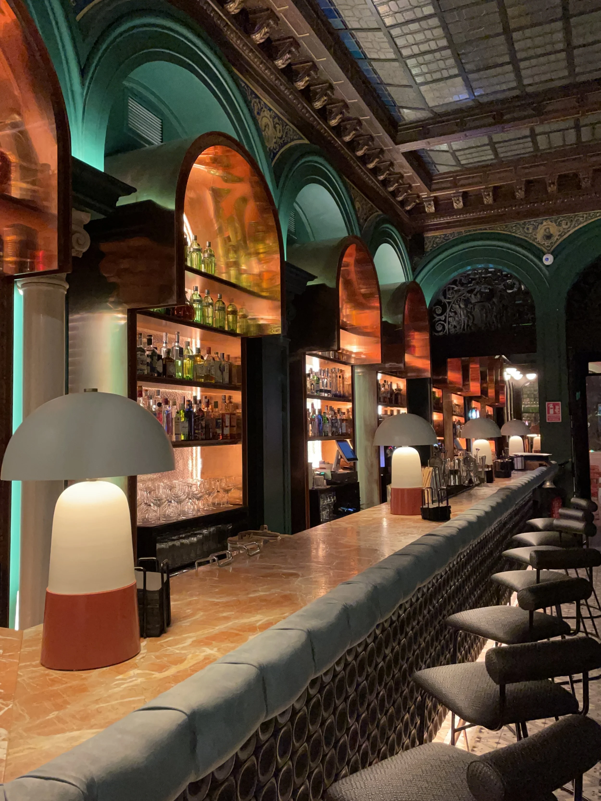  Interior of a bar with a marble countertop, modern lamps, and shelves with various bottles of alcohol behind the bar. The decor includes arched panels with warm lighting, green walls, and ornate ceiling detailing.