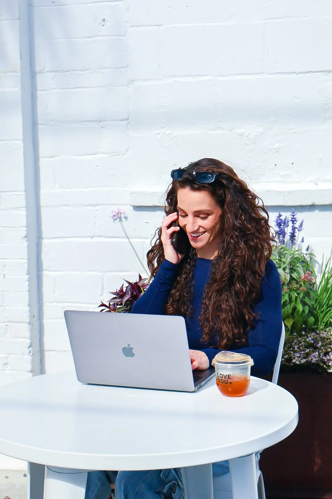 A woman with long curly hair talks on her cellphone while sitting at a white outdoor table with a silver MacBook and a cup of tea, against a white brick wall with flowers in the background.