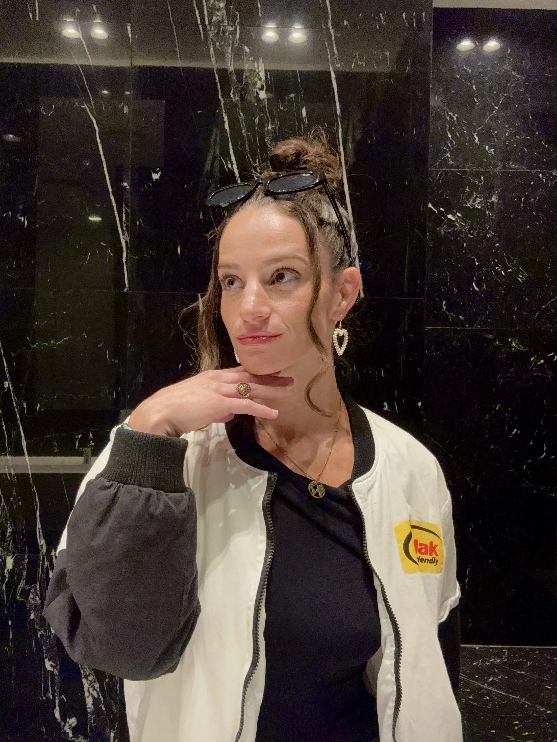 A woman with brown hair tied up in a bun, wearing sunglasses on her head, a white and black jacket with a logo, earrings, a necklace, and a ring, standing against a dark marble wall.