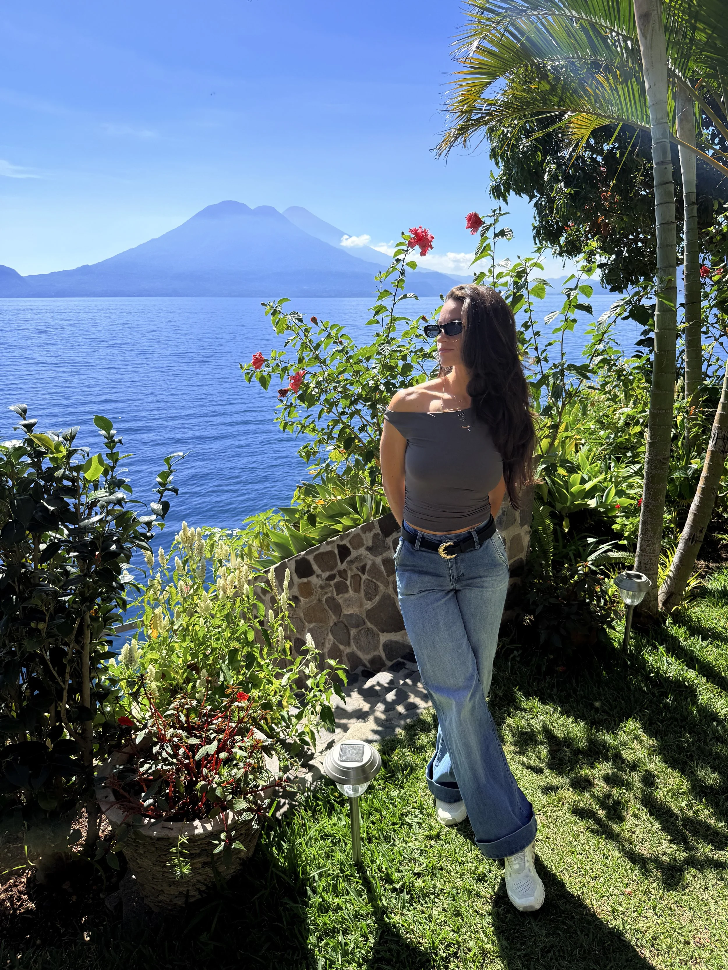A woman with long dark hair, wearing sunglasses, a gray off-the-shoulder top, high-waisted jeans, and white sneakers, standing outdoors near lush green plants and flowers, with a large body of water and a volcano in the background on a sunny day.